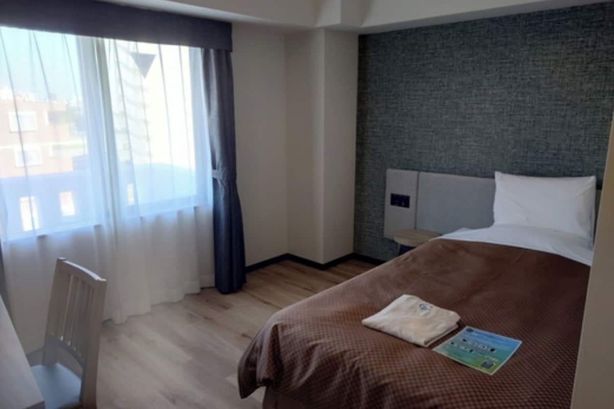 Ein Hotelzimmer mit Bett, Stuhl, einem Fenster mit Vorhängen und Blick auf Gebäude.