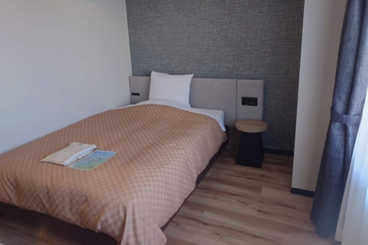 Ein Hotelzimmer mit Bett, Nachttisch, einem kleinen Hocker und einer an der Wand befestigten Bedienkonsole.