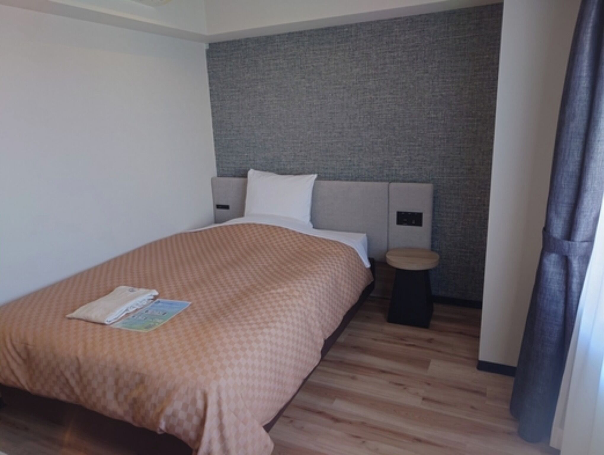 A hotel room with a bed, a bedside table, a small stool, and a wall-mounted control panel.
