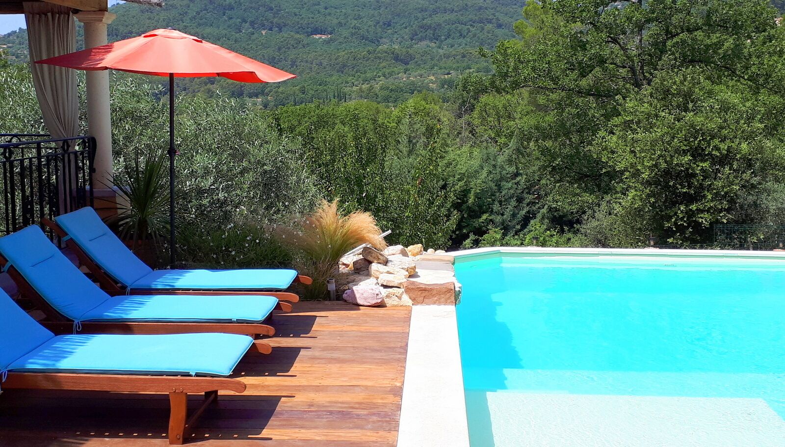A poolside area with wooden loungers and a red umbrella, surrounded by lush greenery and a mountainous backdrop.