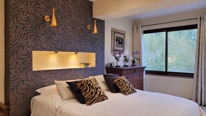 A bedroom with a patterned wallpaper, a bed with zebra-patterned pillows, a bedside table with a camera, and a window with curtains. - Exceptional luxury house, poolhouse area and heated swimming pool (Seillans)