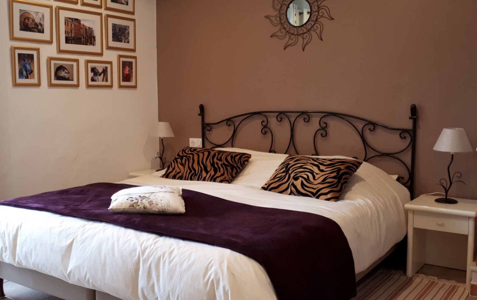 A neatly made bed with a purple blanket and two tiger-striped pillows, a small bedside table with a lamp, and a wall with framed pictures.