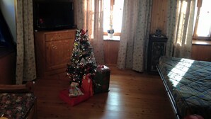 Une pièce chaleureuse aux murs en bois, avec un lit recouvert d’une courtepointe, une télévision, un sapin de Noël et une fenêtre ornée de rideaux.