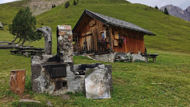 Une cabane rustique en bois, avec des fondations en pierre, entourée d’une colline verdoyante et adossée à une montagne rocheuse.