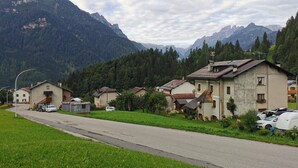 Une route de campagne bordée de maisons et de voitures garées, entourée de montagnes et de verdure.