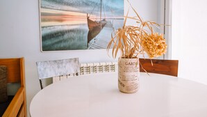 A dining area with a white table, a vase with dried plants, a wooden chair, and a painting of a boat on a pier. - Agi Marina 8 Apartamento for rent in Roses Girona. (Rosas / Roses)