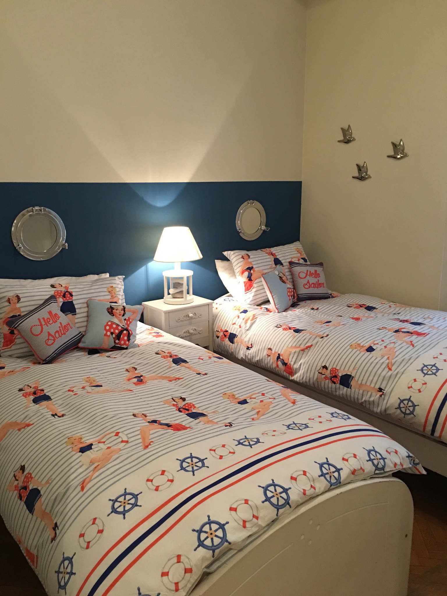 A bedroom with two beds, each with matching bedding featuring nautical-themed designs. There are two bedside tables with lamps, and the room has a blue accent wall with decorative mirrors.
