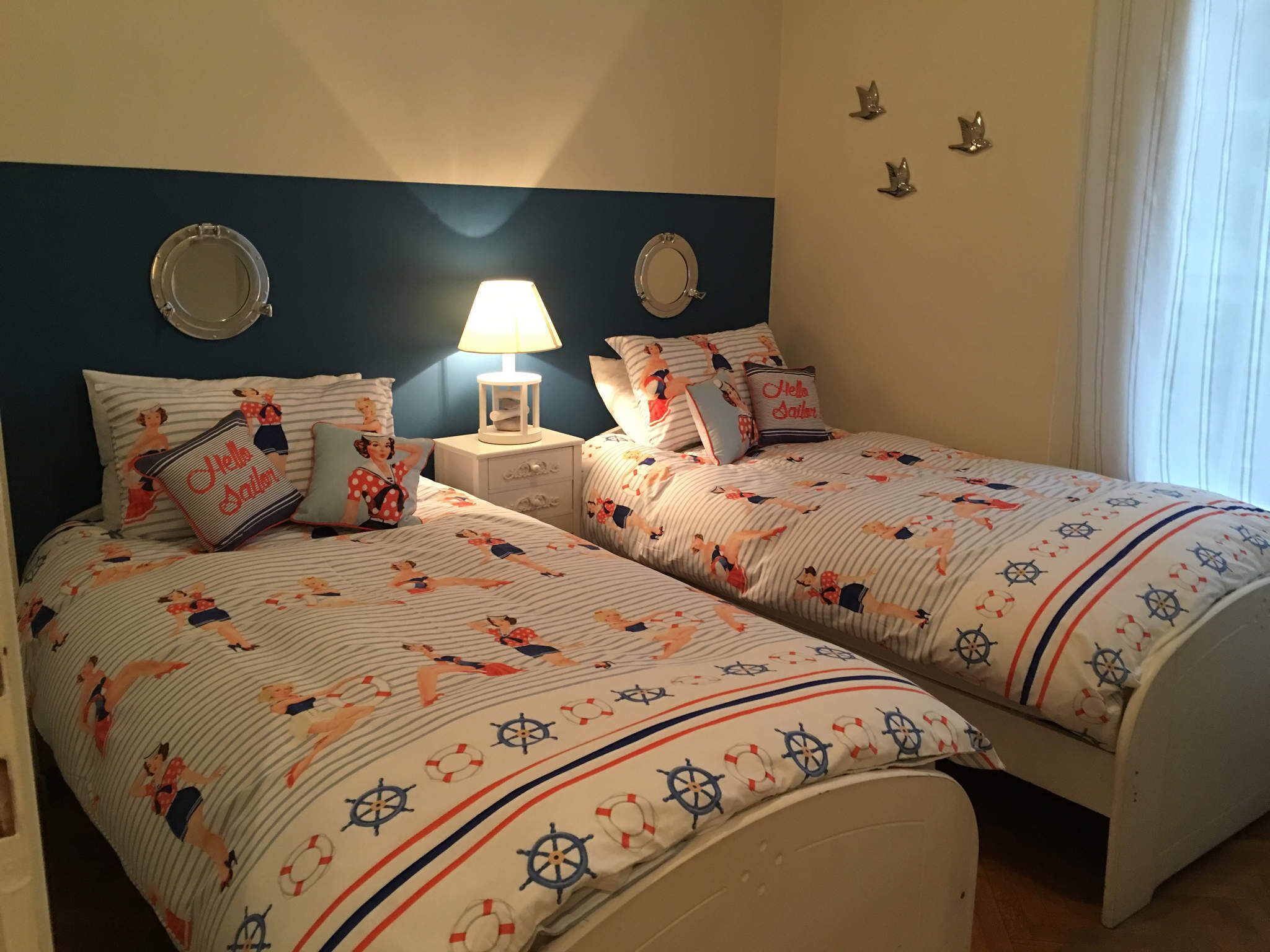 Un dormitorio con dos camas, cada una con ropa de cama a juego con motivos náuticos. Hay una mesita de noche con una lámpara entre las camas, y un espejo montado en la pared sobre cada cama.