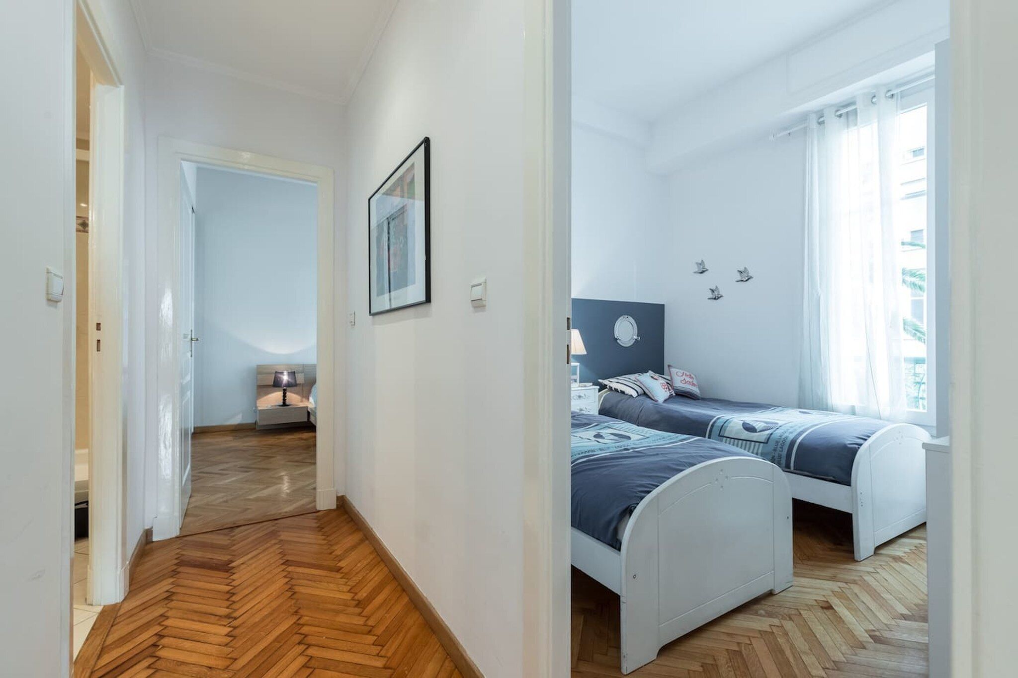 A bedroom with two beds, a wooden floor, a nightstand, a lamp, and a window with curtains.