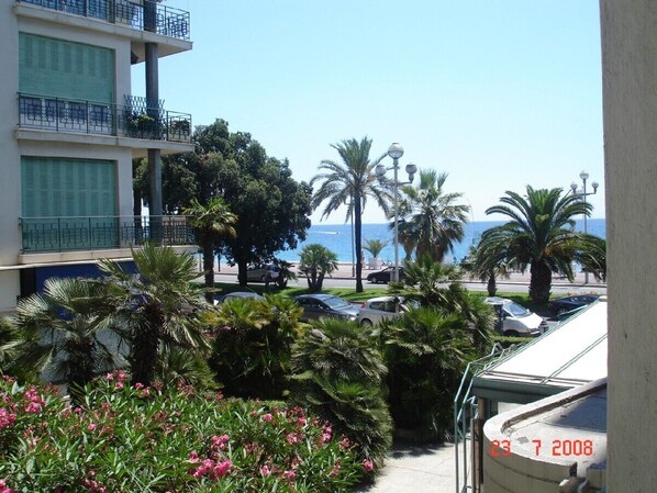 A balcony with a view of the sea, palm trees, and parked cars.