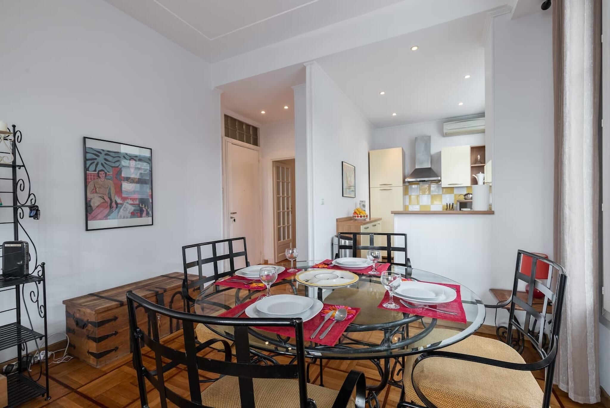 A dining area with a round glass table, black chairs, and red placemats. There is a kitchen in the background with white cabinets and a stove.