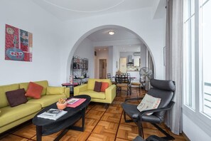 Ein großzügiger Wohnbereich mit einem gelben Sofa, einem schwarzen Sessel und einem Couchtisch. Ein Bogen führt in einen Essbereich mit Tisch und Stühlen.
