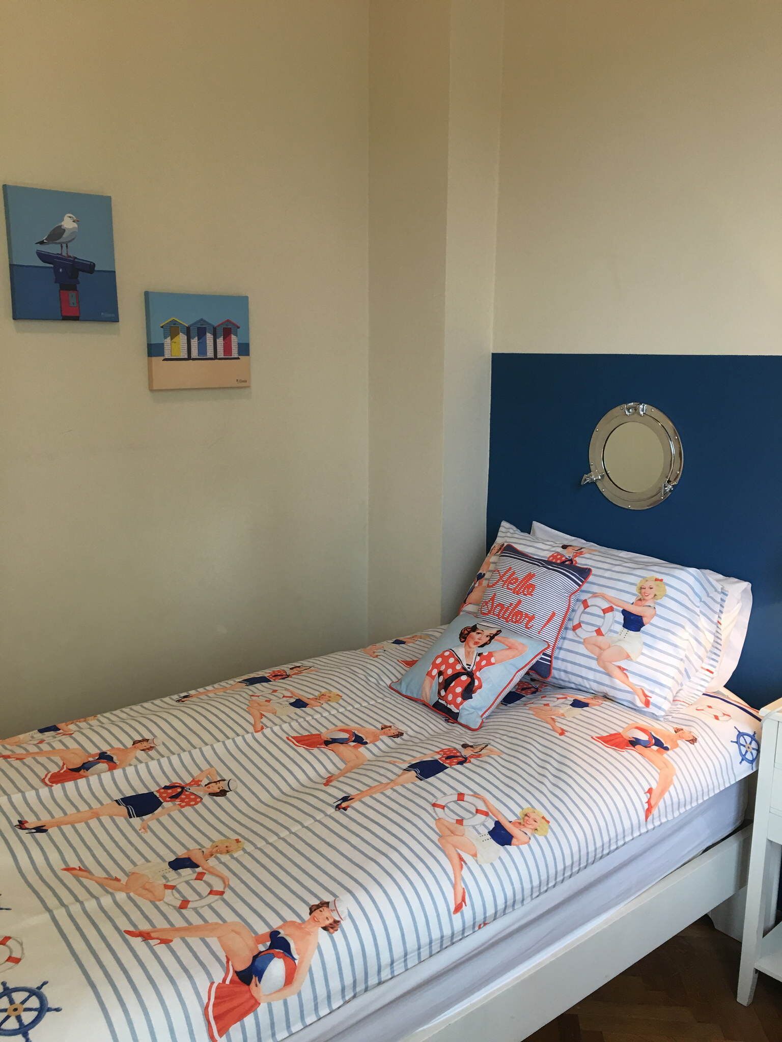 Ein Bett mit gestreifter Bettwäsche und Kissen im Marine-Stil, ein runder Spiegel am Kopfteil sowie zwei Wandbilder mit einer Möwe und bunten Strandhäusern.