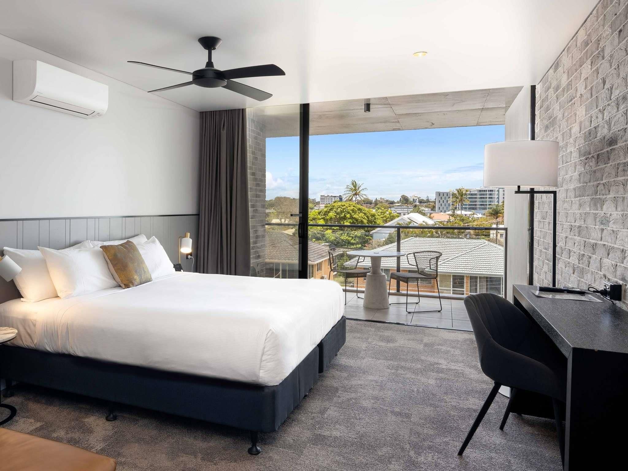 A hotel room with a bed, a desk, a chair, a ceiling fan, and a balcony with a view of the city.