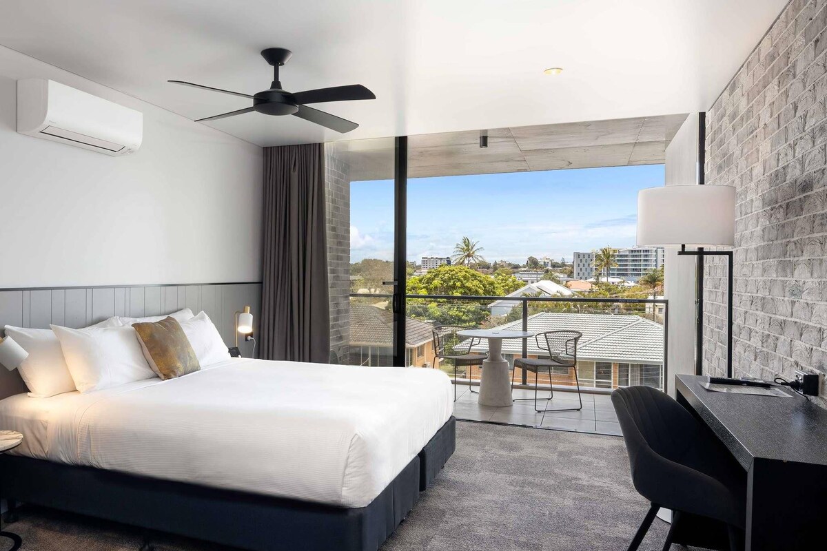 A hotel room with a bed, a desk, a chair, a ceiling fan, and a balcony with a view of the city.