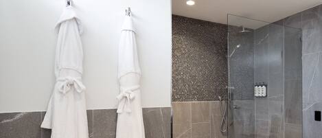A modern bathroom with two white towels hanging on hooks, a wooden bench with towels, and a glass-enclosed shower area with a patterned wall.