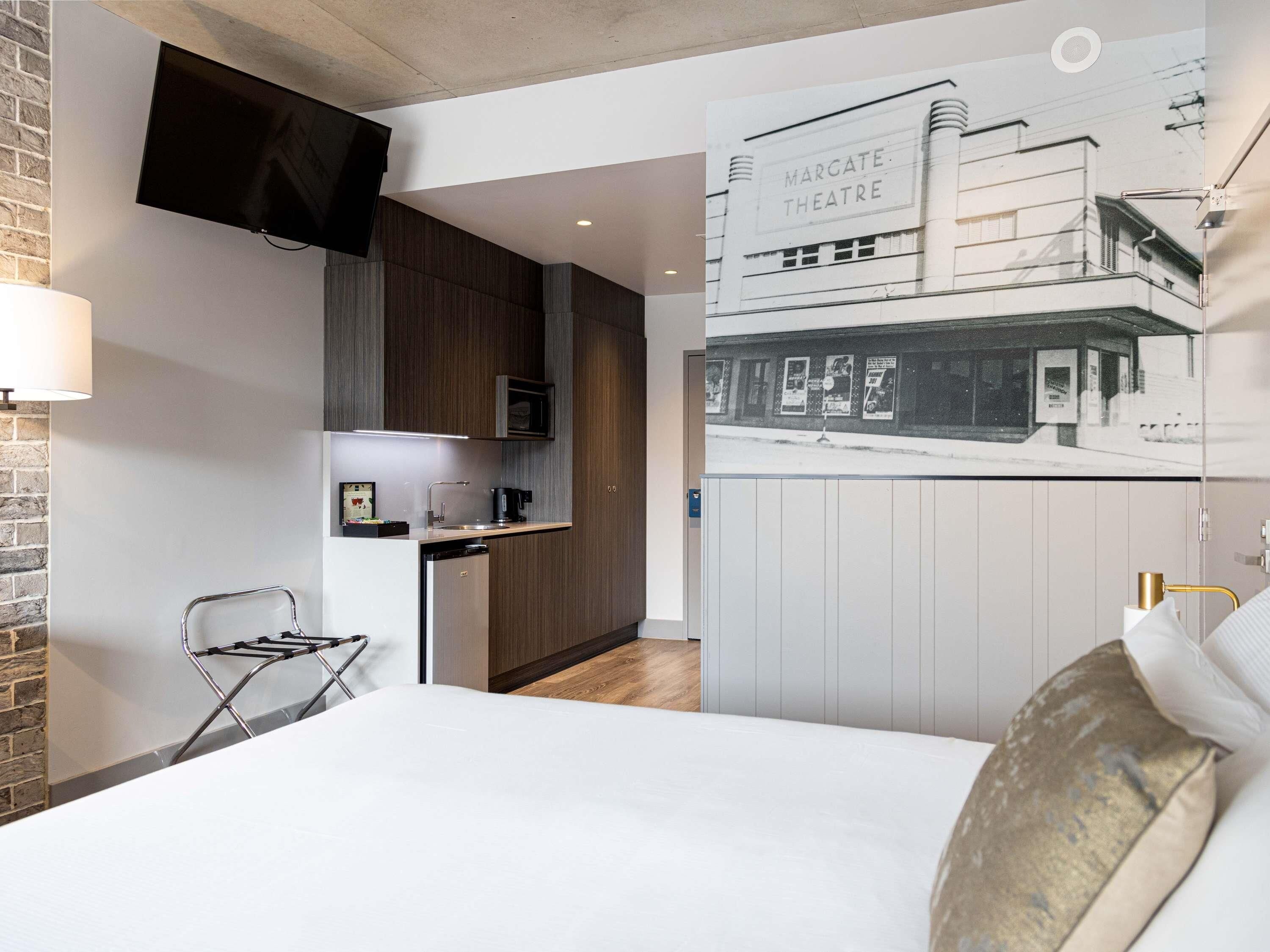 A modern hotel room with a kitchenette, a flat-screen TV, a bed with white linens, and a wall-mounted poster of a theater.