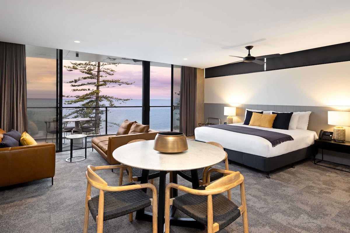 A hotel room with a large bed, a dining table, and chairs. There is a view of the sea and a tree outside the window.
