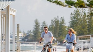 A wooden boardwalk with two people riding bicycles, overlooking a beach and ocean. - The Sebel Brisbane Margate Beach (Margate)