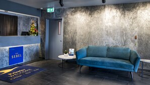A reception area with a blue sofa, a reception desk, a welcome mat, and a wall-mounted plant. - The Sebel Brisbane Margate Beach (Margate)
