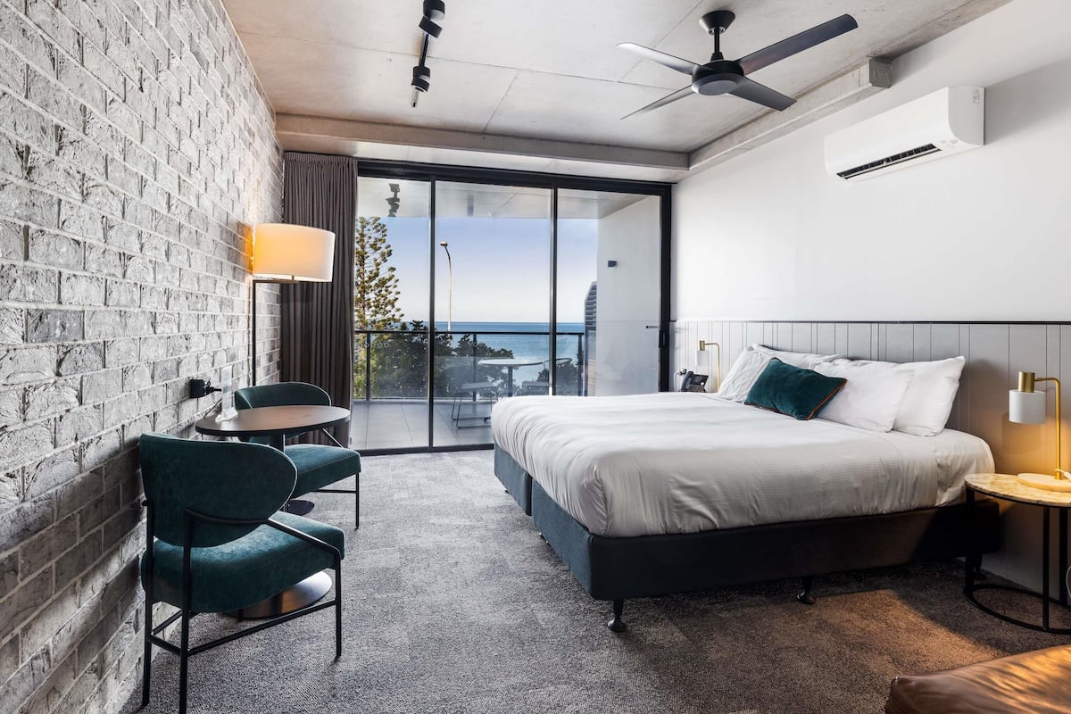 A modern hotel room with a large bed, a balcony with a view, a brick wall, and a ceiling fan.