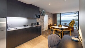 A modern kitchen with dark cabinetry, a stainless steel countertop, and a built-in TV. There is a dining area with a round table and chairs, and a sofa. The room has a large window with a view of the sea.