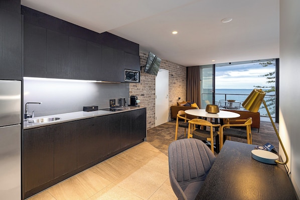 A modern kitchen with dark cabinetry, a stainless steel countertop, and a built-in TV. There is a dining area with a round table and chairs, and a sofa. The room has a large window with a view of the sea. - The Sebel Brisbane Margate Beach (Margate)