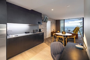 Una cocina moderna con armarios oscuros, encimera de acero inoxidable y televisor integrado. Hay una zona de comedor con una mesa redonda y sillas, además de un sofá. La habitación tiene un ventanal con vistas al mar.