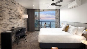 A modern hotel room with a brick wall, a desk with a chair, a bed with white linens, a ceiling fan, and a view of the sea through a balcony door. - The Sebel Brisbane Margate Beach (Margate)