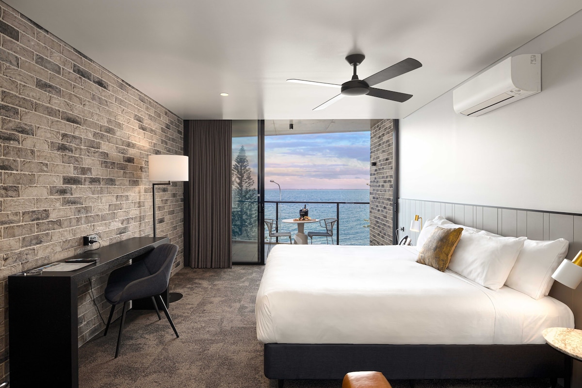 A modern hotel room with a brick wall, a desk with a chair, a bed with white linens, a ceiling fan, and a view of the sea through a balcony door.