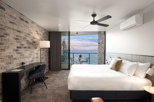 Una habitación de hotel moderna con pared de ladrillo, un escritorio con silla, cama con sábanas blancas, ventilador de techo y vista al mar a través de una puerta de balcón.