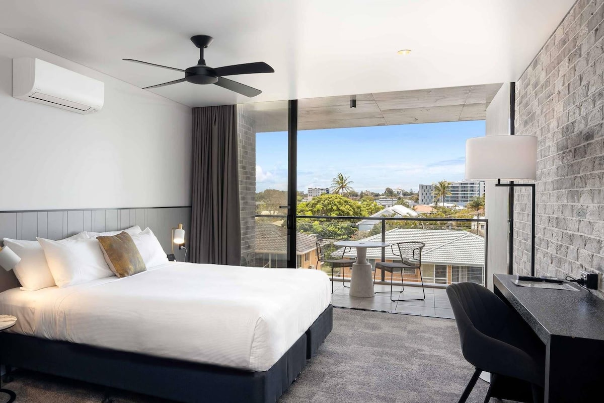A hotel room with a bed, a desk, a chair, a ceiling fan, and a balcony with a view of the city.