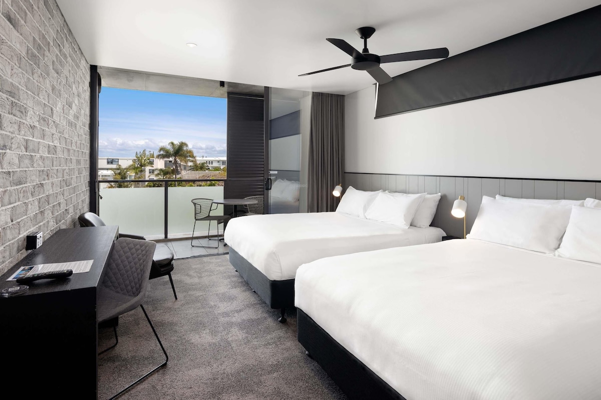 A modern hotel room with two beds, a desk, a chair, a ceiling fan, and a balcony with a view.