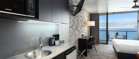 A modern hotel room with a kitchen area, a bed, a desk, and a view of the sea.