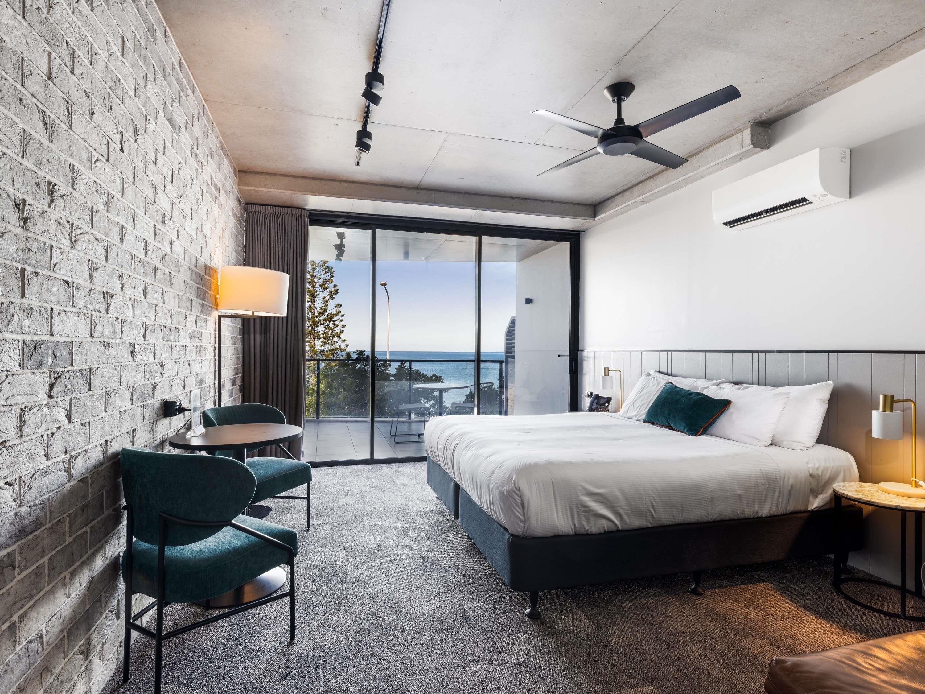 A modern hotel room with a large bed, a balcony with a view, a brick wall, and a ceiling fan.