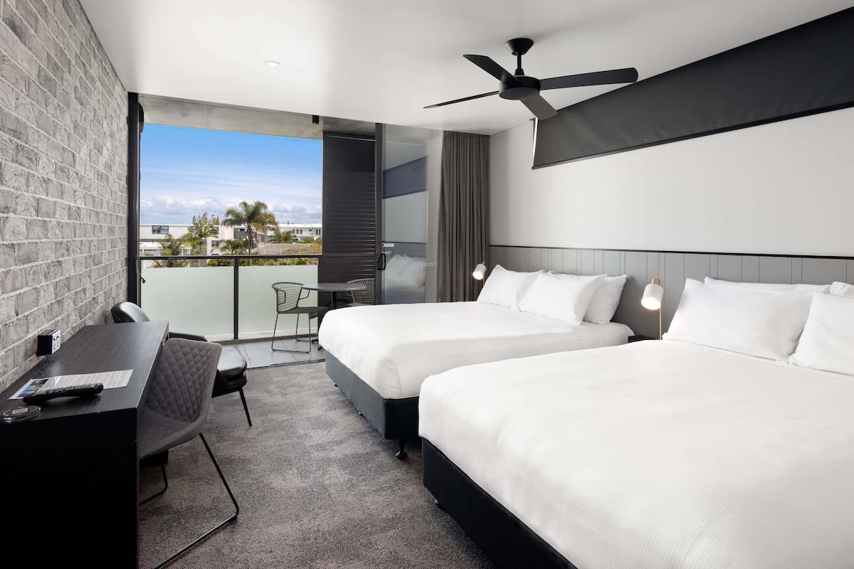A modern hotel room with two beds, a desk, a chair, a ceiling fan, and a balcony with a view.