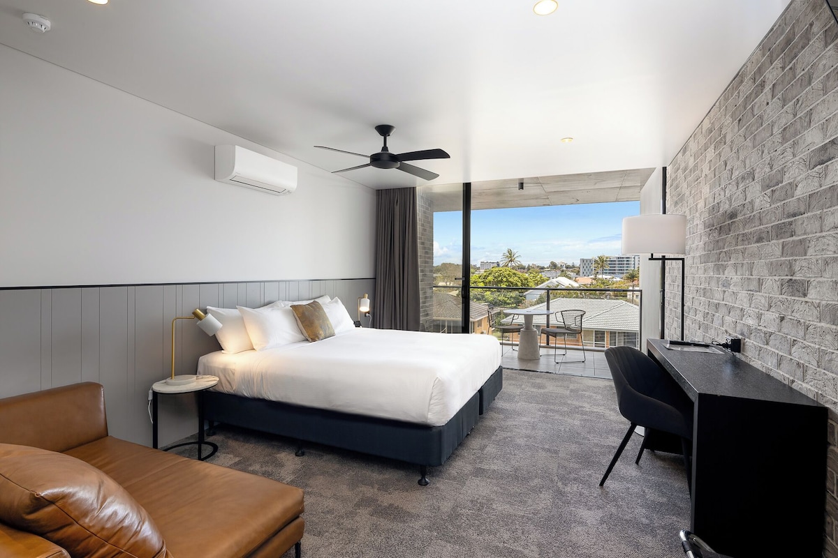 A modern hotel room with a large bed, a desk, and a balcony with a city view.