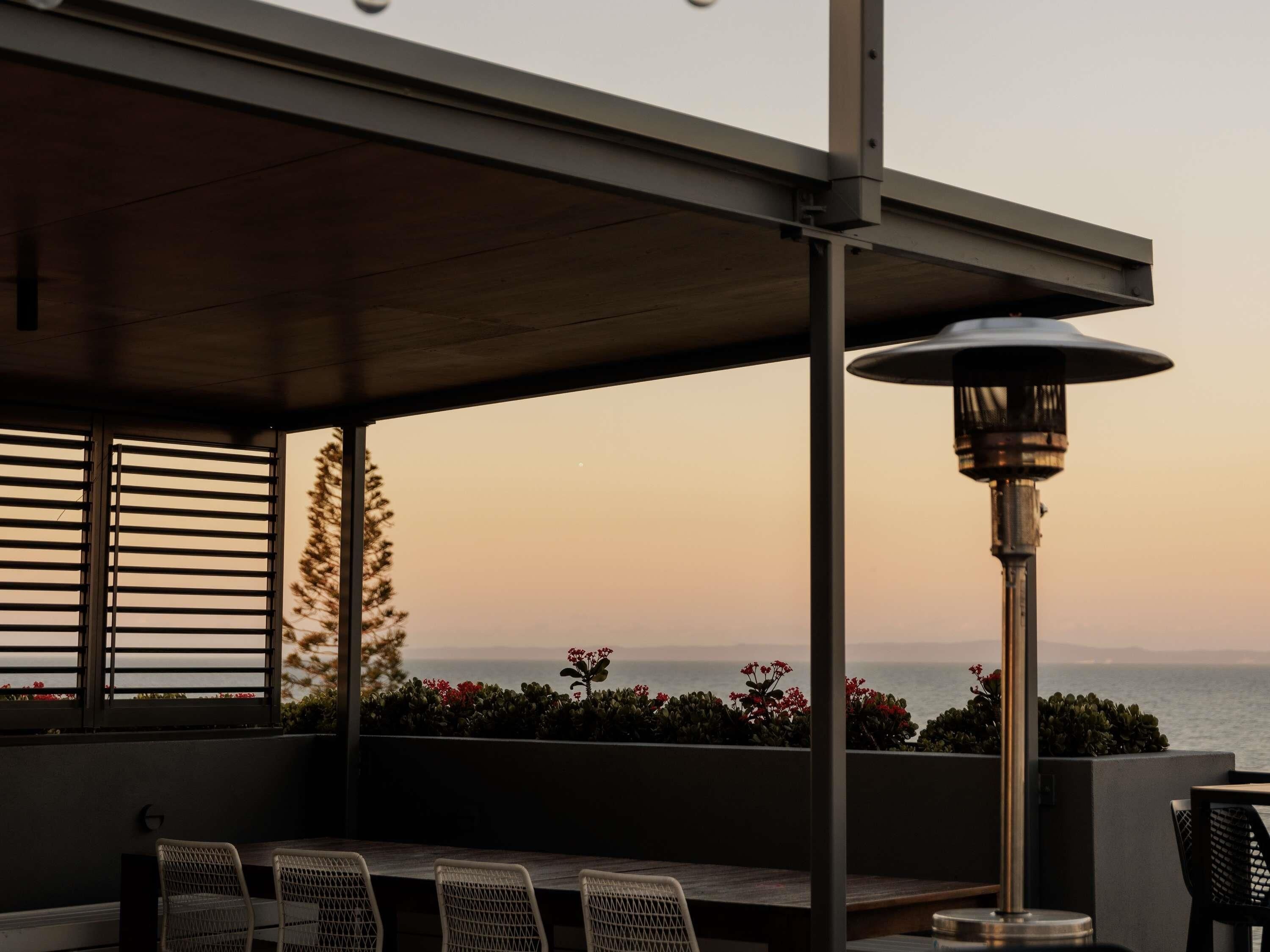 An outdoor patio with a table and chairs, a heater, and a view of the ocean at sunset.