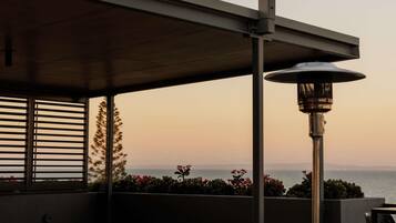 An outdoor patio with a table and chairs, a heater, and a view of the ocean at sunset.