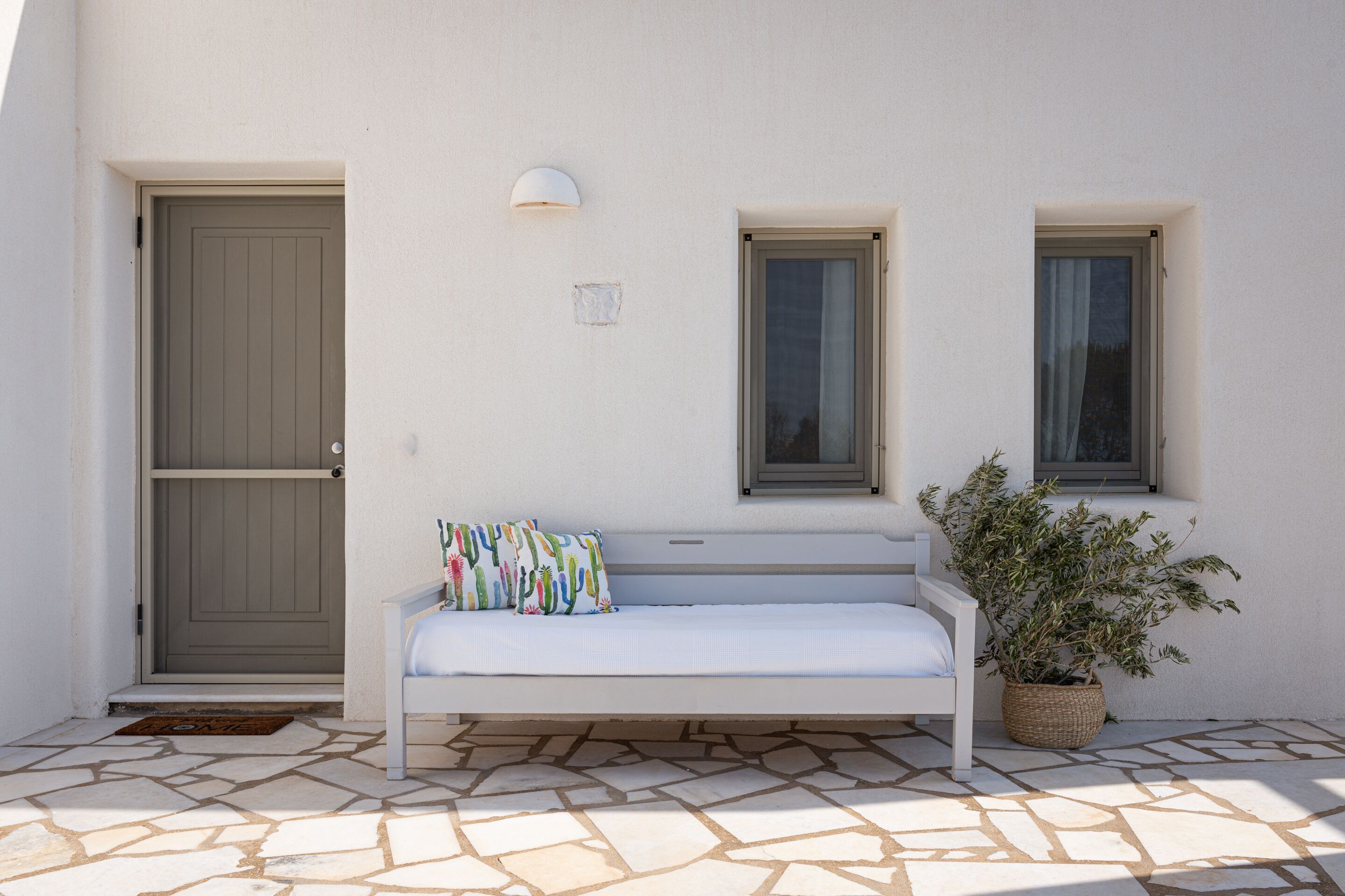 A white bench with a colorful cushion, a potted plant, and a door with a window above it.