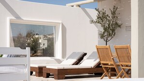Un espace extérieur ensoleillé, aménagé avec des chaises longues en bois et un canapé blanc, un petit arbre, et une vue sur une ville côtière, visible à travers une grande fenêtre.