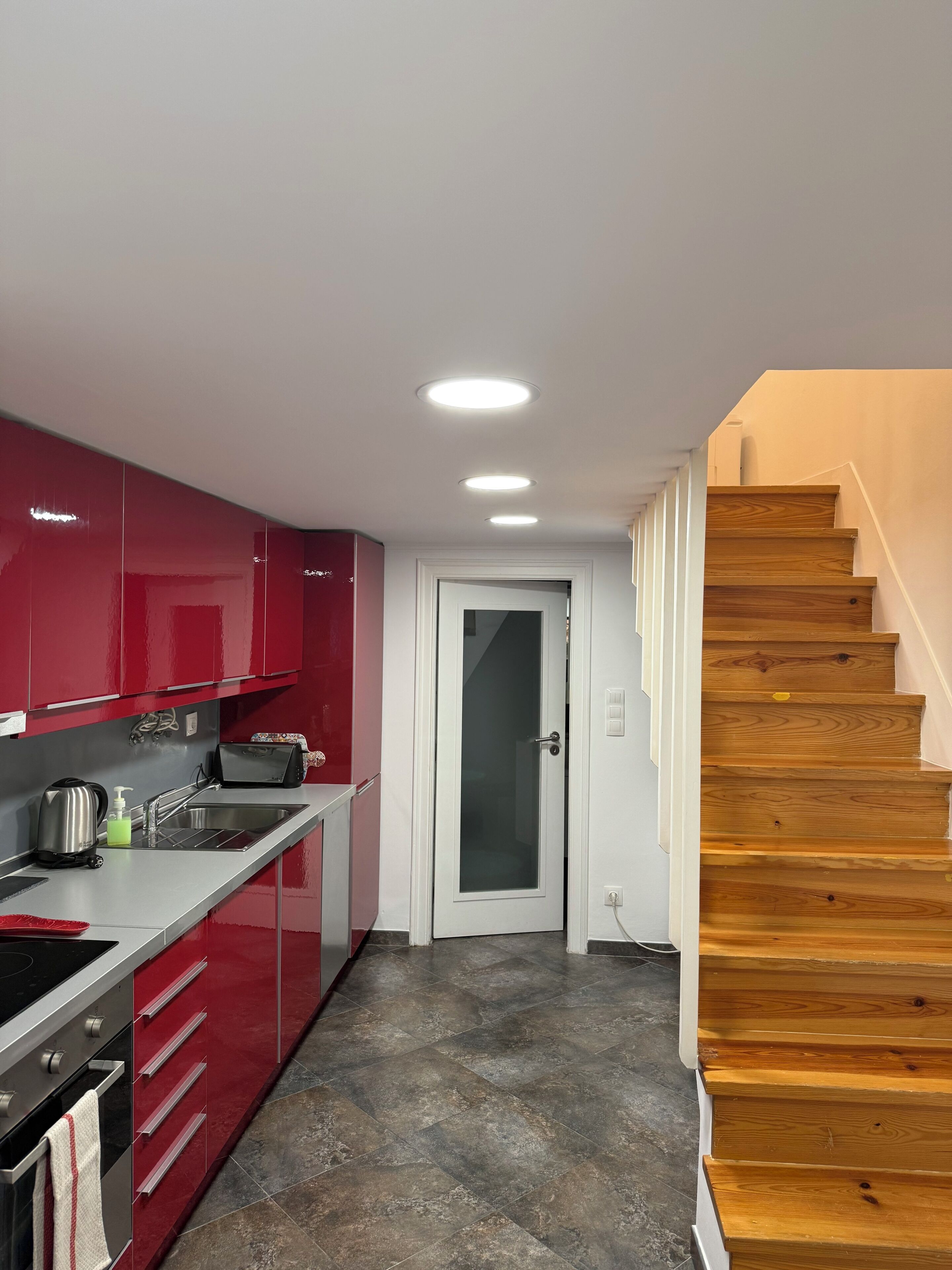A modern kitchen with red cabinets, a stainless steel sink, and a wooden staircase.