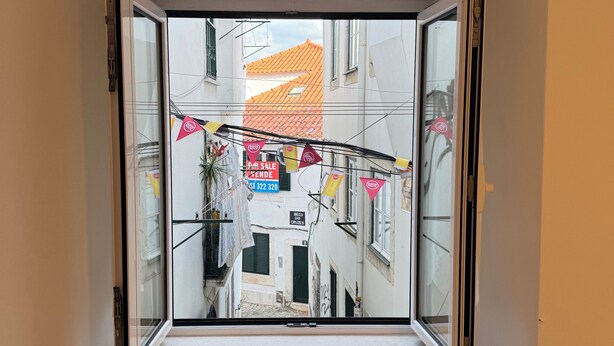 A room with a window seat, a view of a narrow street with buildings, and a 'For Sale' sign. - Romantic Alfama - Duplex Apartment in Lisbon, Alfama (Lisboa)