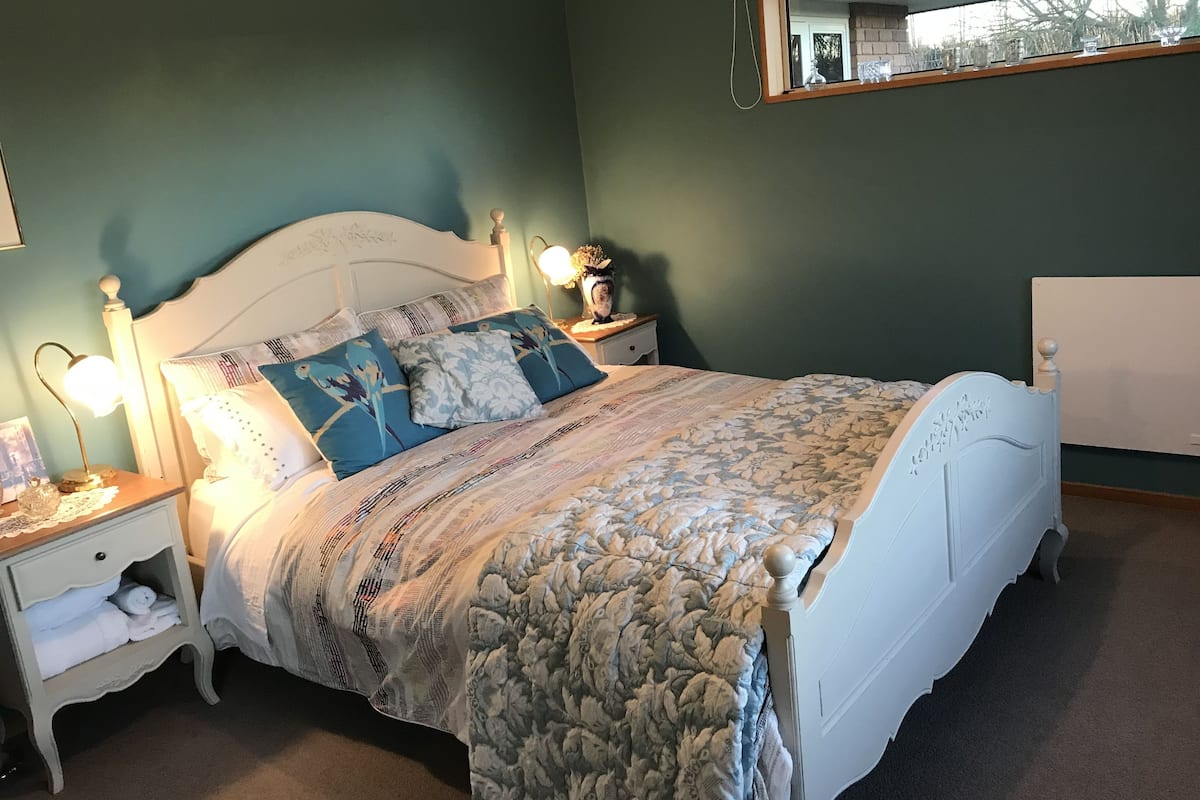 Cama bem arrumada com edredom floral e várias almofadas, luminária de cabeceira e mesa de cabeceira com abajur.