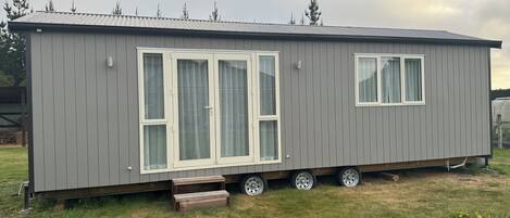 A grey mobile home with two large sliding doors and a smaller window, parked on a grassy area with trees in the background.