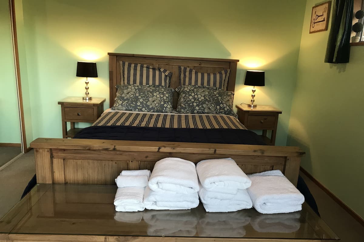 Uma cama bem arrumada com roupa de cama listrada e almofadas florais, com duas mesas de cabeceira com abajures de cada lado, e uma mesa de vidro com toalhas dobradas na frente.