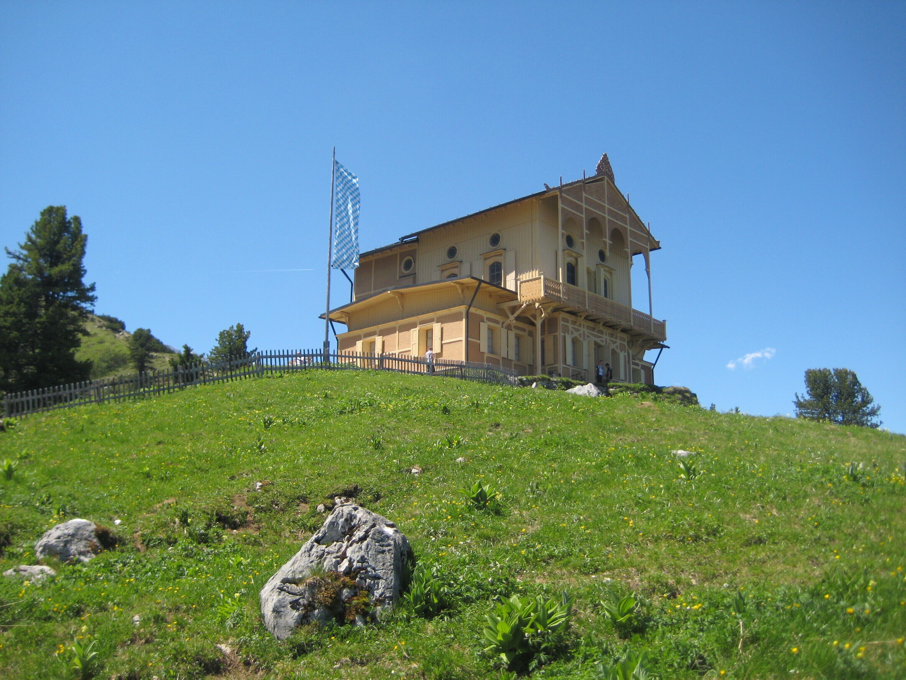 A large, two-story building with a balcony on the upper floor, situated on a grassy hill with a clear blue sky.