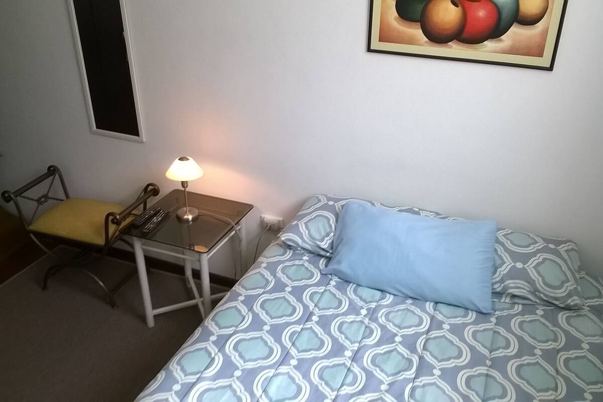 A small room with a bed, a table with a lamp, a chair, and a wall-mounted picture.