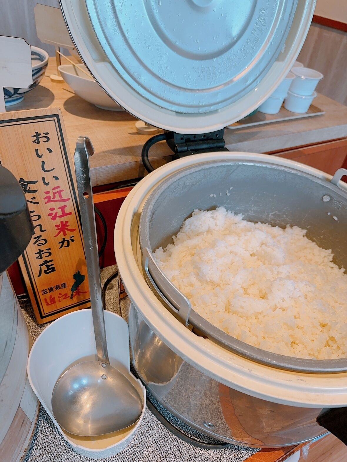 Een keuken met een rijstkoker vol witte rijst, een opscheplepel en een houten bordje met Japanse tekst.