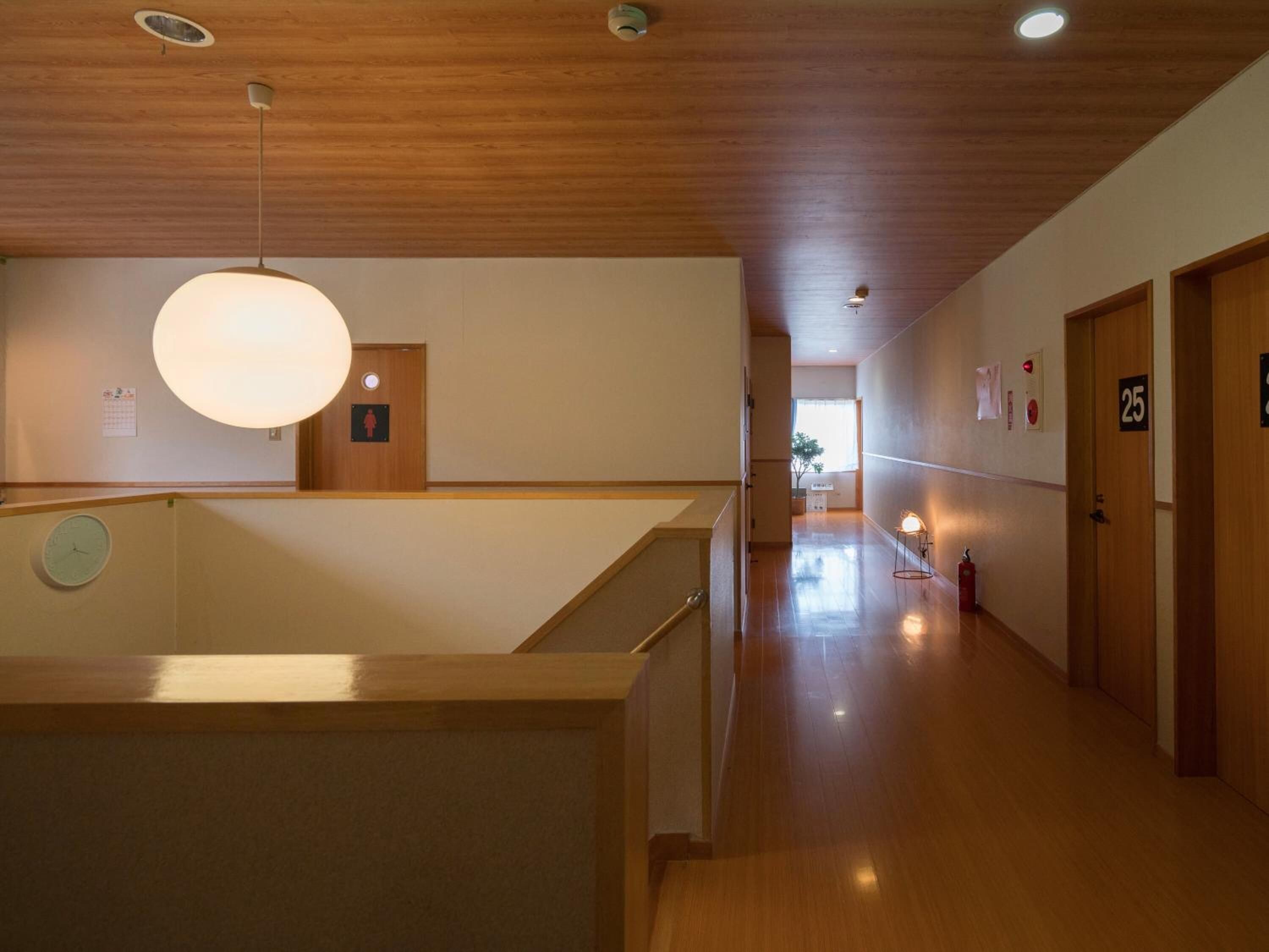 Un couloir d’hôtel avec un plancher en bois, un comptoir de réception et un grand lustre suspendu. On y trouve les portes numérotées 25 et 26, ainsi qu’un extincteur fixé au mur.